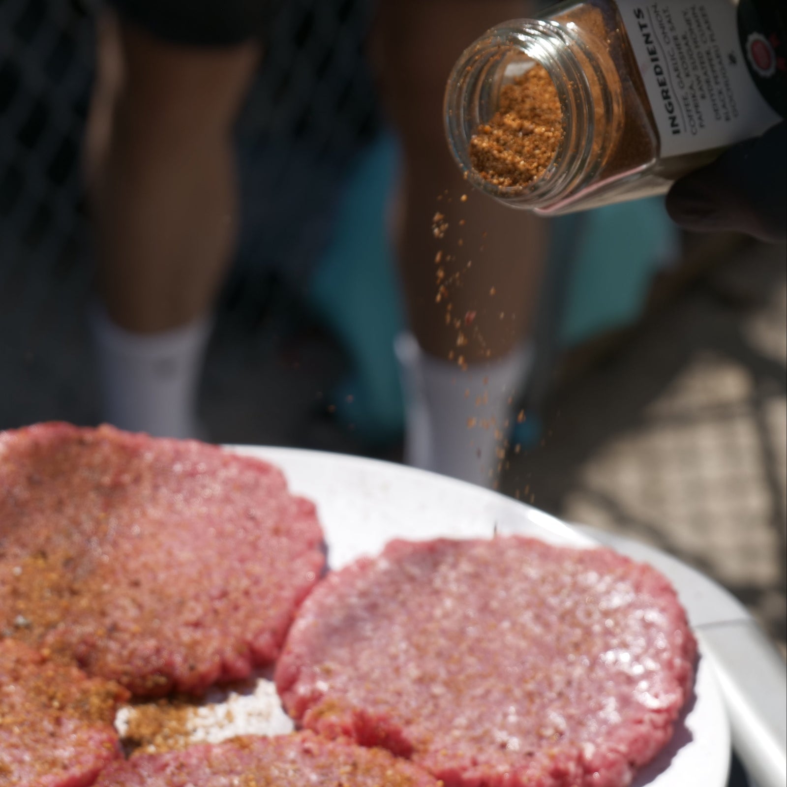 Uncooked burger patties on a plate with a person seasoning them outdoors.